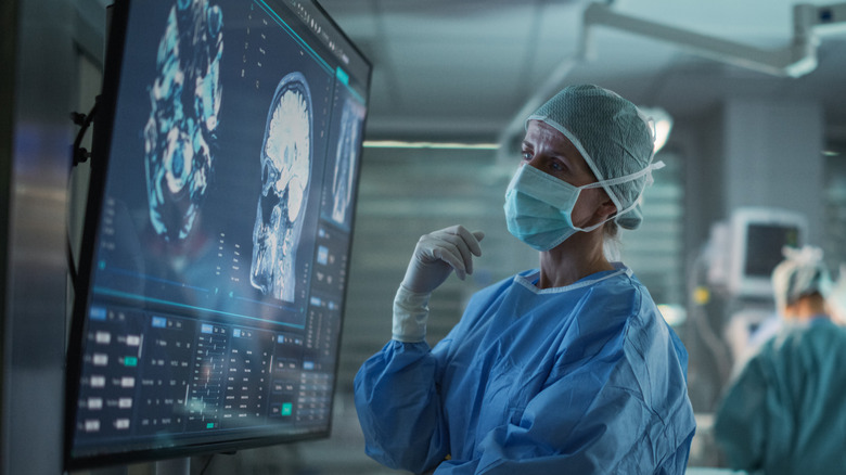 A masked surgeon looks at a brain scan while other medical staff attend to a patient in background in blue-lit image