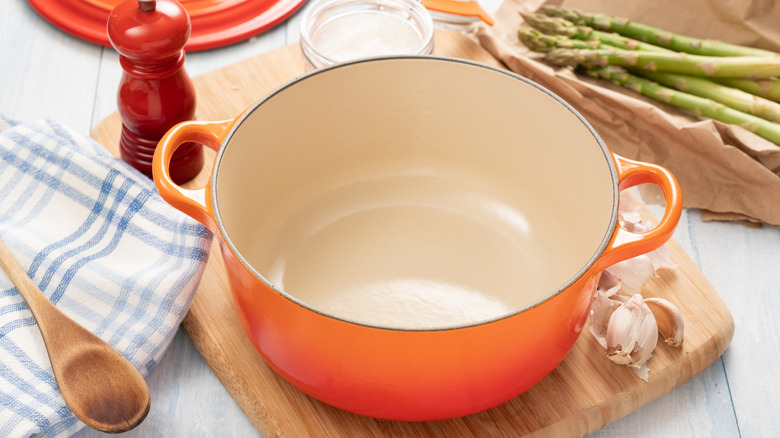 A warm orange Le Creuset style dutch oven in classic orange, open to reveal enameled cast iron inside, set on a table full of garlic, asparagus, spoon and towel