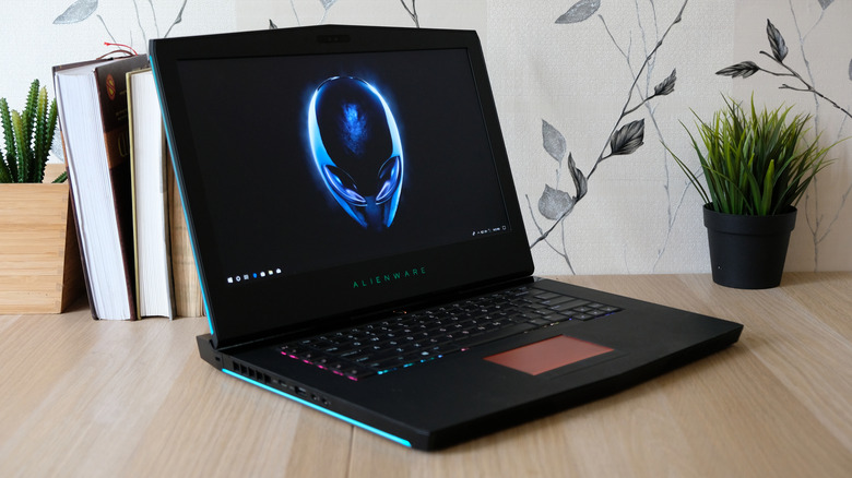 An Alienware laptop open and powered on sits on a woodern desk next to some cacti and books