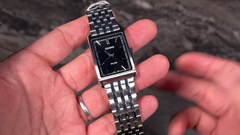Person holding a rectangular Casio MTP-B215 watch with a black dial and silver multi-link metal bracelet