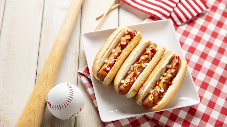 Three hot dogs on a white plate on a white wooden picnic bench.
