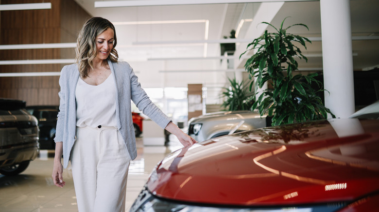 A woman looking fondly at a car parked inside a car dealership with her hand on the hood.