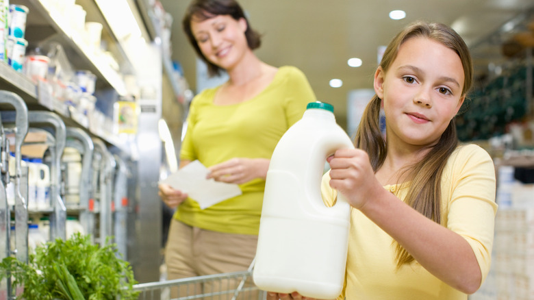 A little girl holding up a jug of milk in a grocery store with her mother smiling in the background.