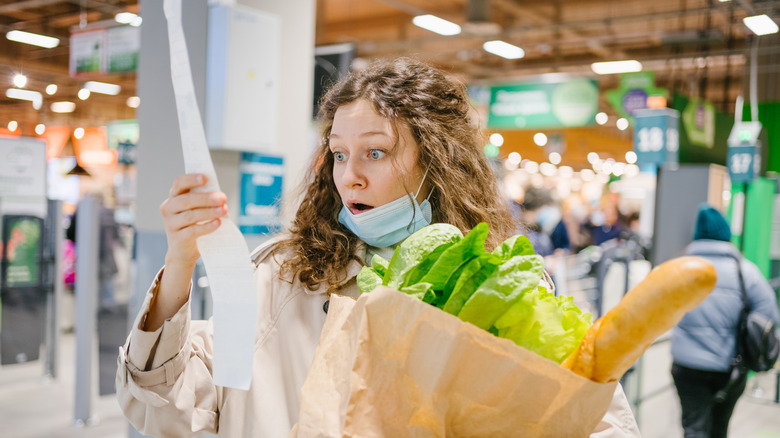 A young woman with a shocked expression looking at her grocery bill while holding a grocery bag in a grocery store.
