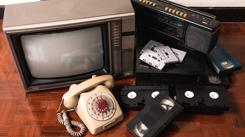 An old console TV, rotary phone, VHS tapes and VHS player, and cassette tapes and a tape player on a table.