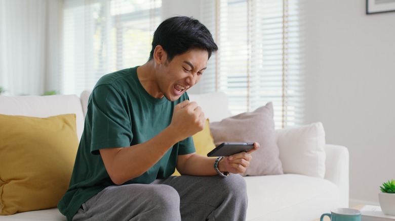 A young man relax sitting easy at home on a couch excited while staring at something on his phone.
