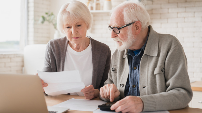 A mature couple look worriedly at a bill while sitting at a kitchen table with a computer open, during the day