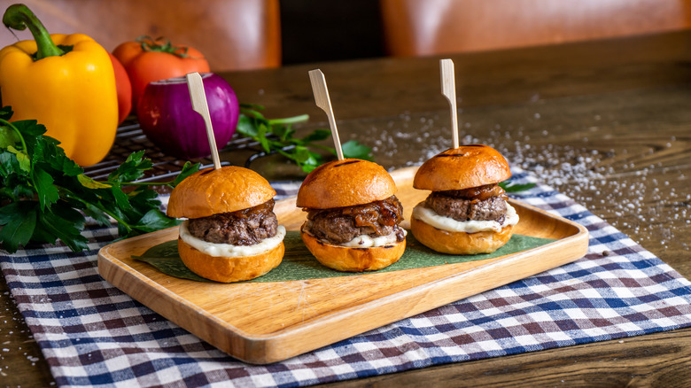 Three mini beef burgers on a small tray in front of fresh peppers and onion