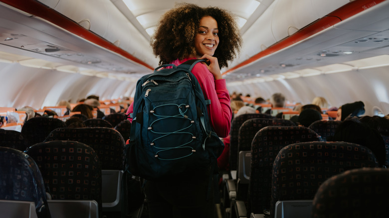A smiling woman with a backpack looking back in an airplane aisle