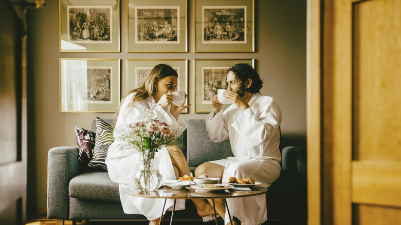 A young couple sitting in a hotel suite sipping coffee in bathrobes
