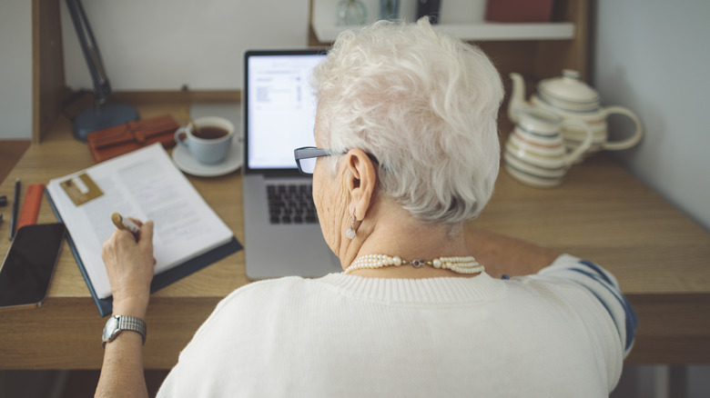 A senior woman at home writing something in a ledger while sitting in front of a laptop