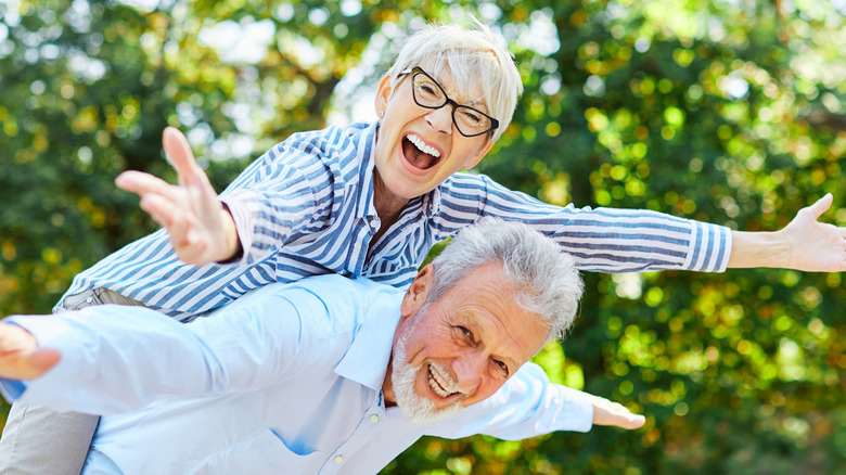 A smiling senior couple enjoying the outdoors with the woman laying on the man's back and both people spreading out their arms.