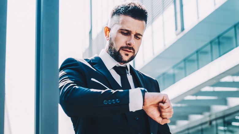 Thoughtful businessman in elegant suit standing on city street checking the time on his watch.