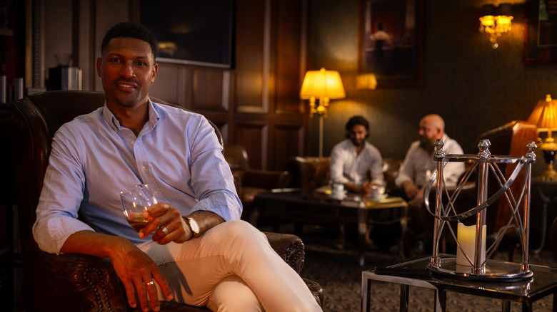 A well dressed man seated in a private club holding a glass of whiskey and smiling.