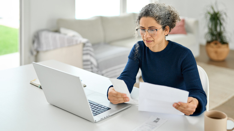 Mature woman reading receipts in front of laptop.