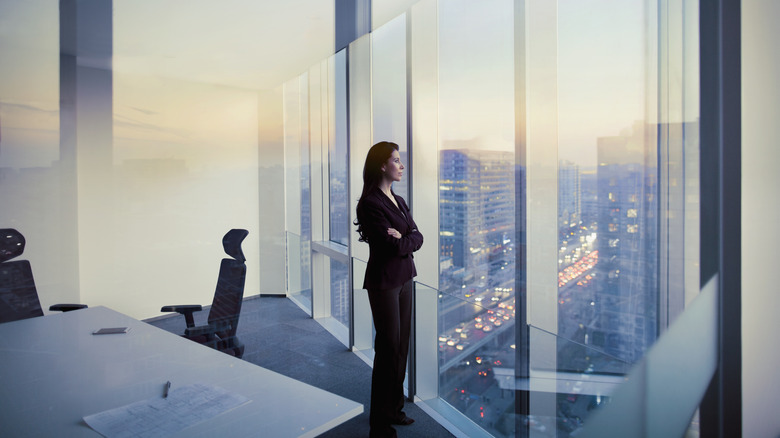 A female CEO staring out the window of a large corner office at s sprawling city.