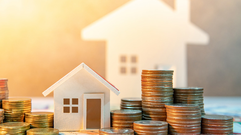 A small paper house on a table surrounded by coins with a larger blurred paper home cutout in the background.