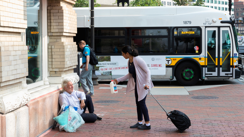 Woman offers milk to man sitting on street as bus goes by