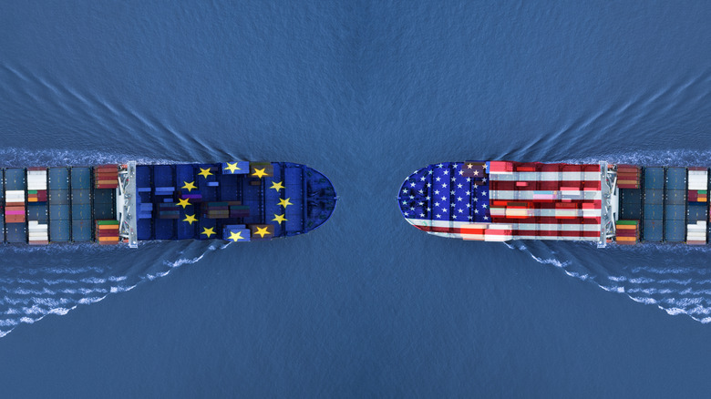 An overhead view of two cargo ships, one bearing the EU flag and the other the USA stars and stripes, sail at each other in a collision course.