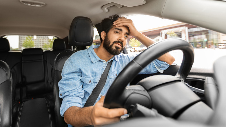 A man sitting in a car's driver's seat with his hand on his forehead looking unhappy.
