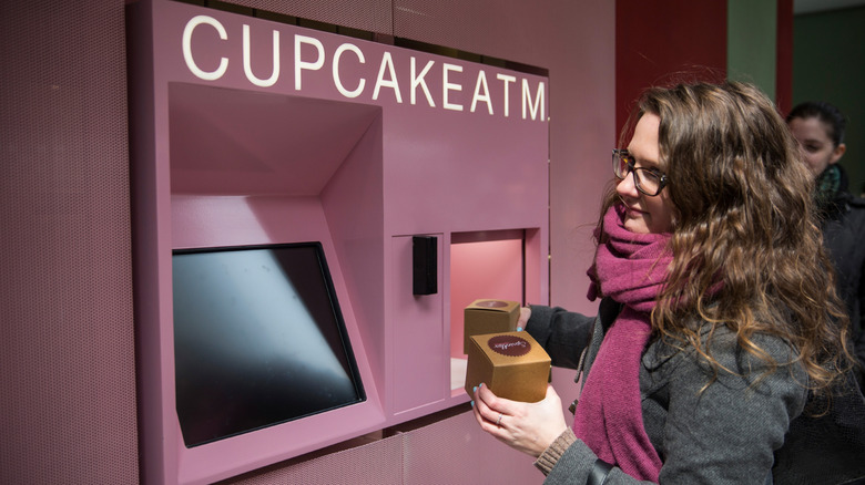 A woman holding cupcake boxes while standing in front of a pink cupcake ATM.