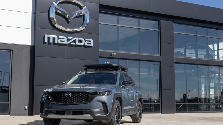 A black Mazda CX-50 sits on the pavement outside of a Mazda dealership on a sunny day.
