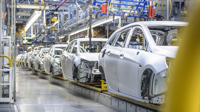 A line of unpainted, unfinished sedans roll through an assembly line in a brightly-lit automotive plant.