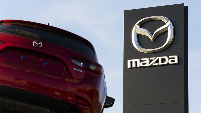 A red Mazda vehicle on left side of frame, a large black and silver Mazda dealership sign on right side of frame, taken during a clear blue day.