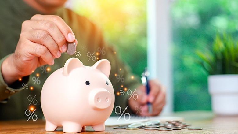 A hand dropping a coin into a pink piggybank with a pile of coins sitting next to the piggybank on a wooden tabletop