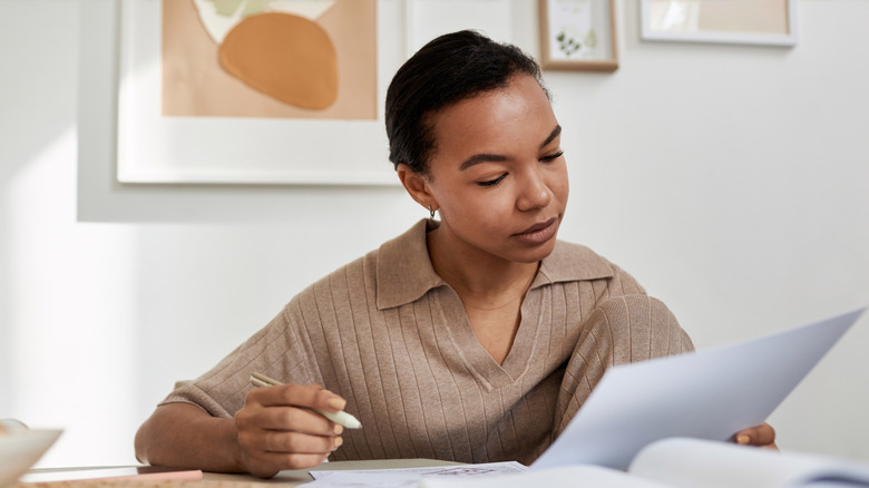 A young black woman with short hair, wearing a tan ribbed polo shirt, sits at a table with books on it, staring intently at a document held on her left hand while holding a pen on her right hand