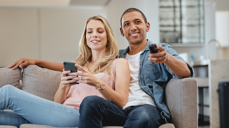 A man changes the TV channel while a woman dials a number on her phone