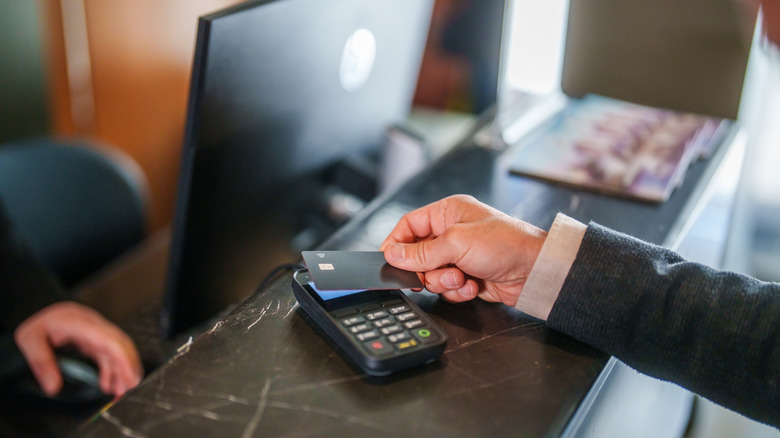 A close-up of a man's hand holding a credit card over a credit card machine at a hotel desk with a computer monitor in the background.