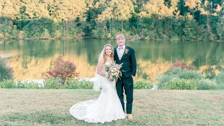A smiling bride and groom posing in front of a lake surrounded by trees.