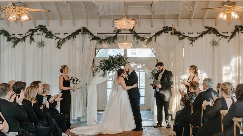 A newly married couple kissing in a wedding venue while guests applaud.