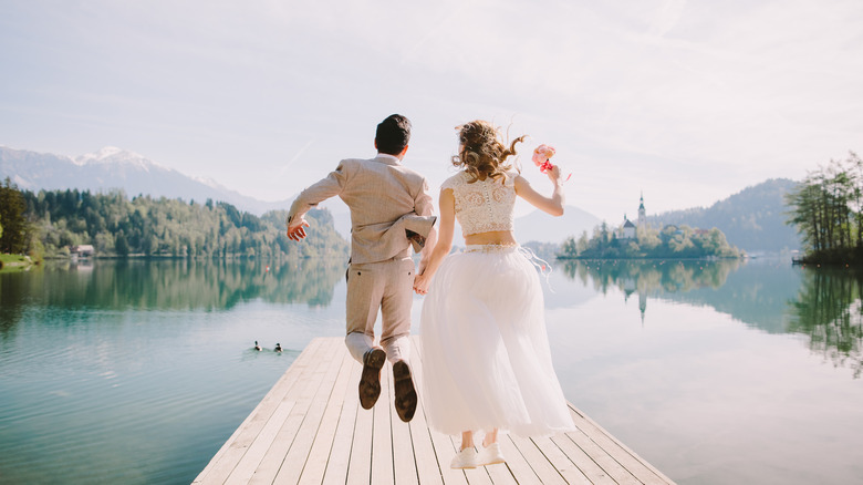 A bride and groom hopping on a pier overlooking a lake and trees.