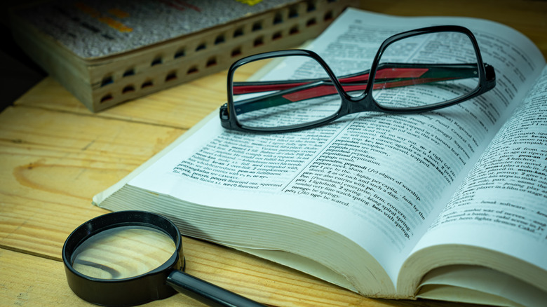 Open dictionary on a wooden table with reading glasses and magnifying glass.