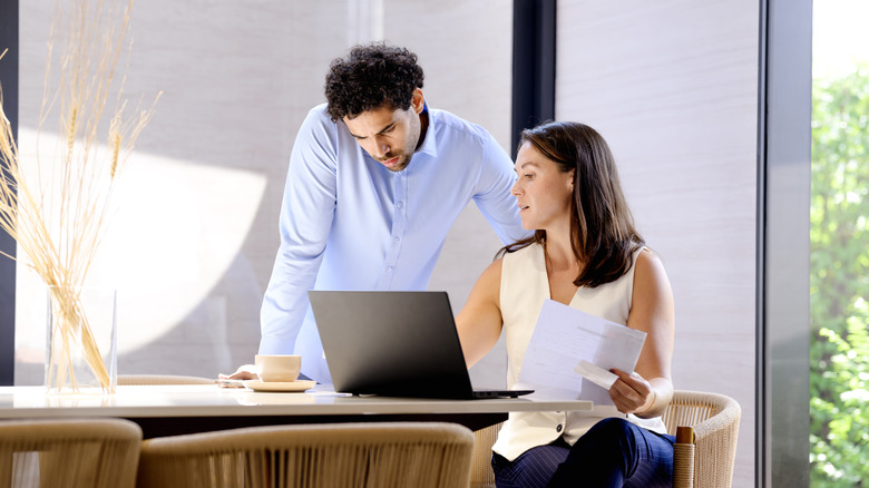 A man standing over a woman who is sitting while looking into a laptop