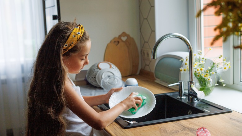 A little girl washing dishes over a sink