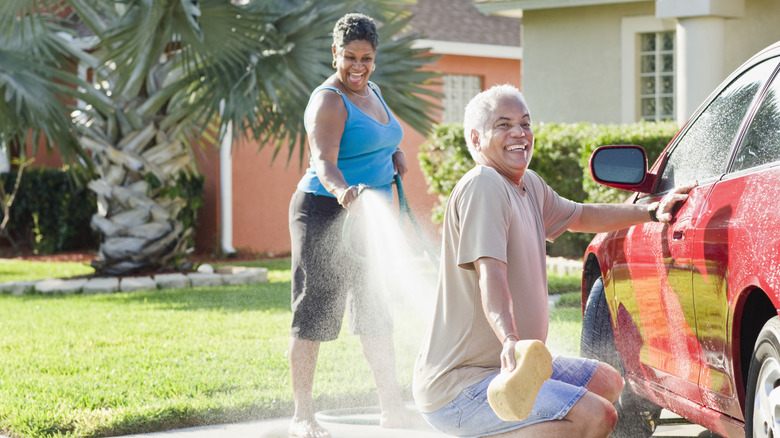A family washing a car outside while a woman playfully hose down a man