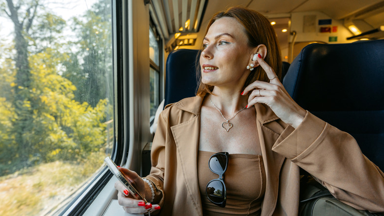A Gen X woman with earbuds holding her phone while gazing out the window of a moving train.