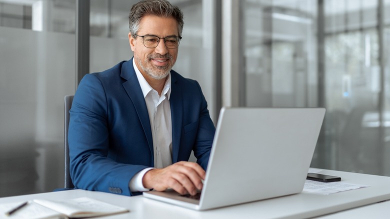 A man in a blue suit sitting at a desk working at a laptop
