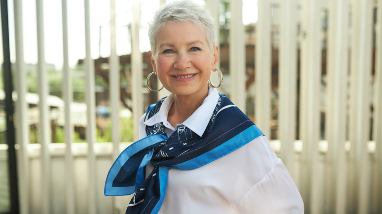 A smiling mature woman in professional attire standing in front of office windows.