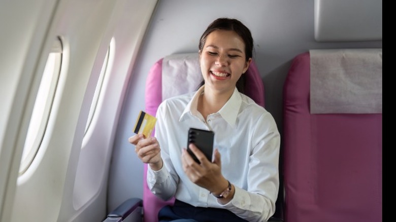 A young woman sitting on a plane and smiling while paying for something on her phone with a credit card.