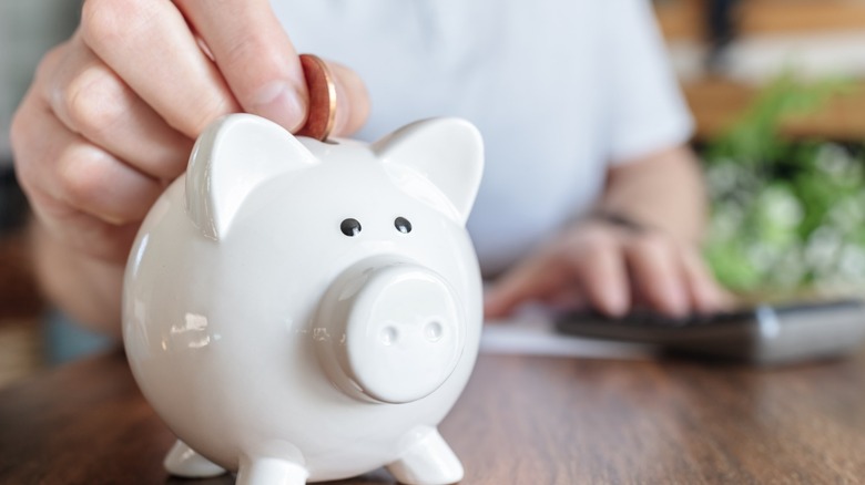 Hand placing a coin into a white piggy bank, with a blurred calculator and greenery in the background