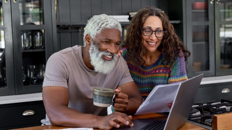 Smiling older couple reviewing documents together at a kitchen table while using a laptop, one holding a coffee mug