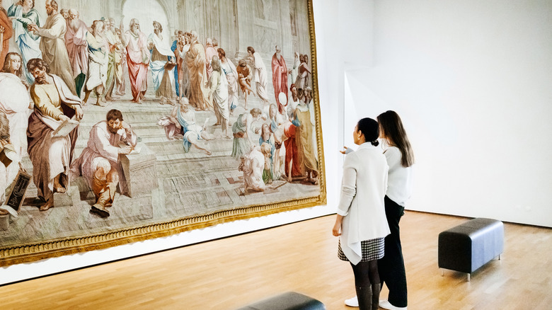 Two women stand in front of a large, detailed historical painting in an art gallery.
