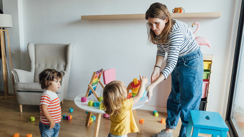 Positive young female babysitter in casual clothes playing with infant children in cozy playroom with colorful educational toys.