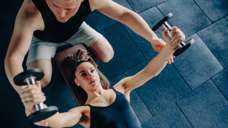 A dedicated male personal trainer acts as a coach, guiding a woman in lifting a dumbbell at the gym.