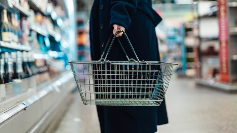 Cropped shot of young person carrying shopping basket, standing at produce aisle in supermarket.