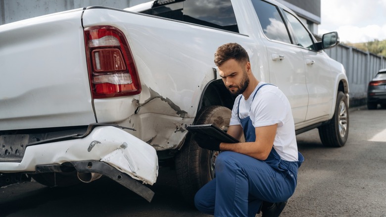 Mechanic kneeling beside a white pickup truck with a heavily dented rear bumper and quarter panel, inspecting damage on a tablet